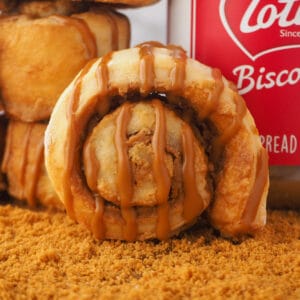 Biscoff Cinnamon Rolls 15 Close up Biscoff scroll on a board with cookie crumbs, with a stack of scrolls and BIscoff jar behind it.