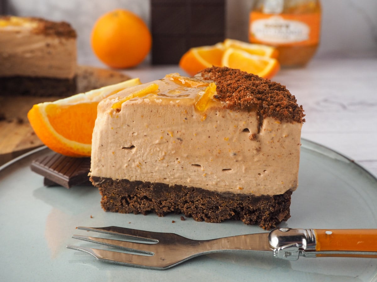 Slice of cheesecake with fork, rest of cake, fresh oranges and dark chocolate in the background.