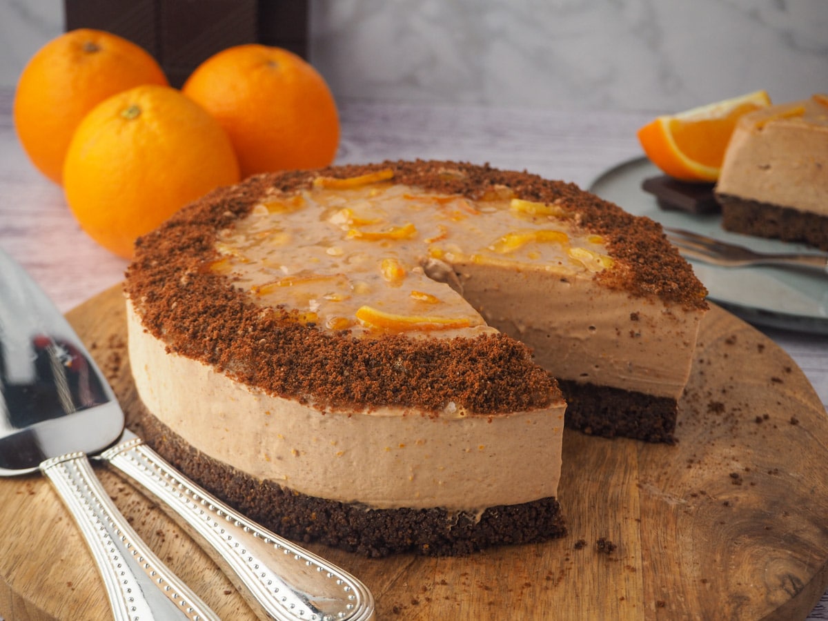 Chocolate Orange Cheesecake 17 Serving cake, with silverware and slice of cake and fresh orange and chocolate in the background.