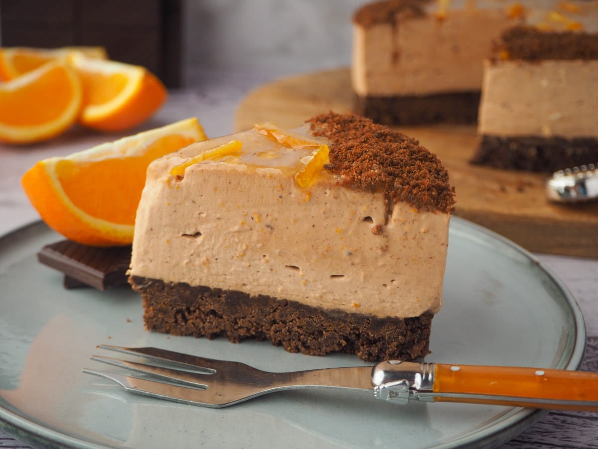 Slice of cheesecake with fork, rest of cake, fresh oranges and dark chocolate in the background.