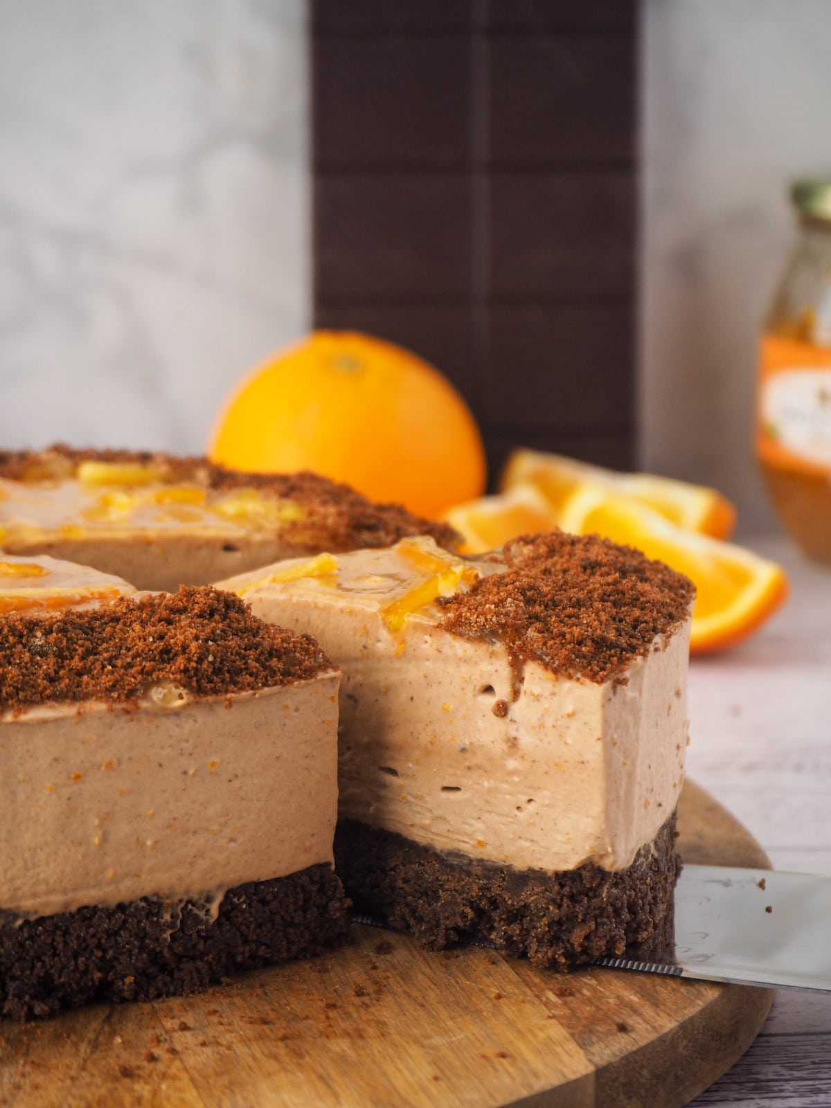 Serving a slice of cheesecake with fresh oranges and dark chocolate in the background.