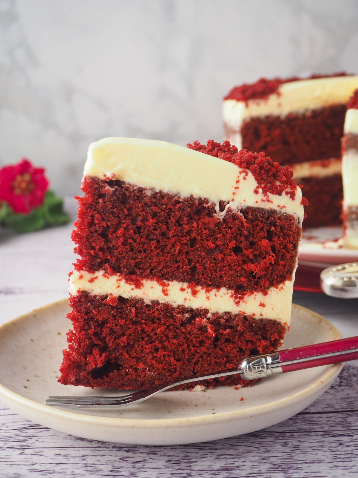 Slice of cake on a plate with fork and cake and flowers in the background.