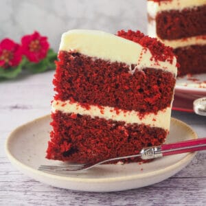 Chocolate Red Velvet Cake 25 Close up slice of chocolate red velvet cake on a plate with fork and cake and flowers in the background.