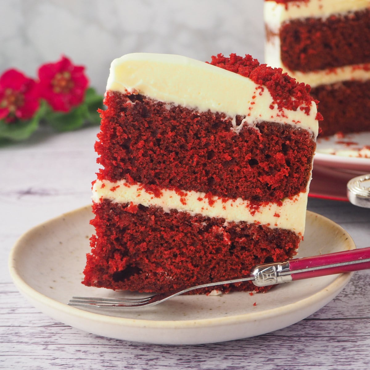 Close up slice of chocolate red velvet cake on a plate with fork and cake and flowers in the background.