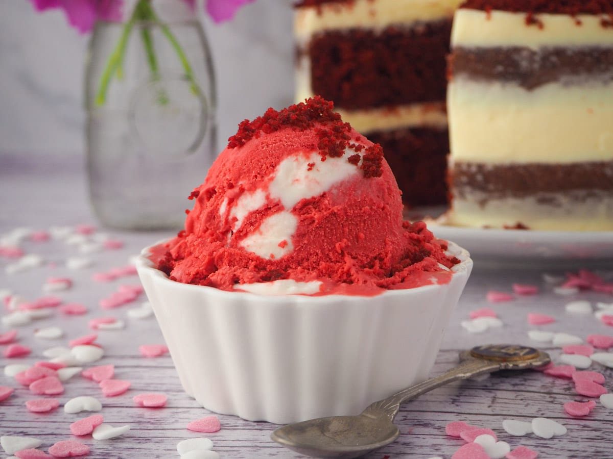 Scoop of ice cream with spoon and heart sprinkles, with red velvet cake and pink flowers in the background.