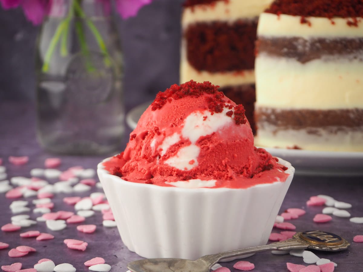 Scoop of ice cream with spoon and heart sprinkles, with red velvet cake and pink flowers in the background.