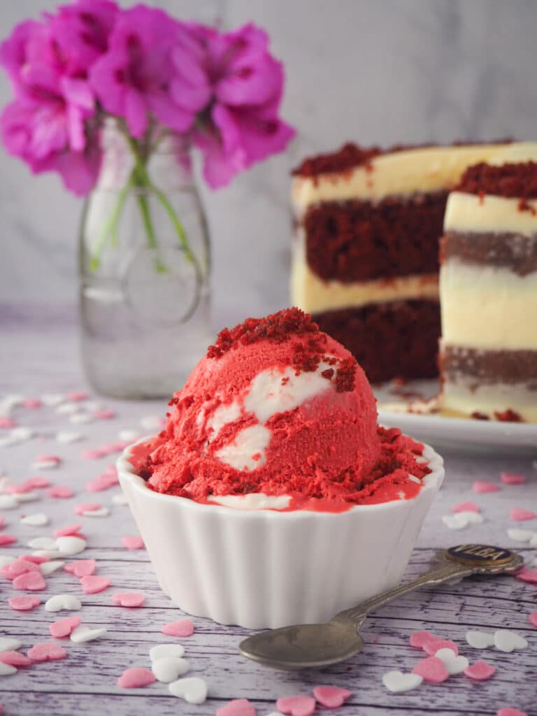 Scoop of ice cream with spoon and heart sprinkles, with red velvet cake and pink flowers in the background.