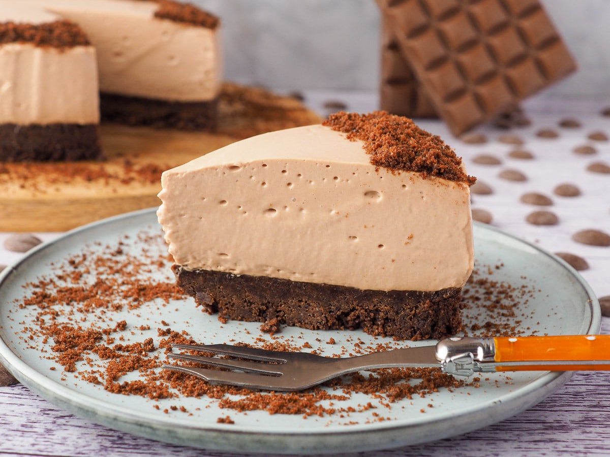 Slice of cheesecake on a plate with a fork and rest of cheesecake and block of chocolate in the background.
