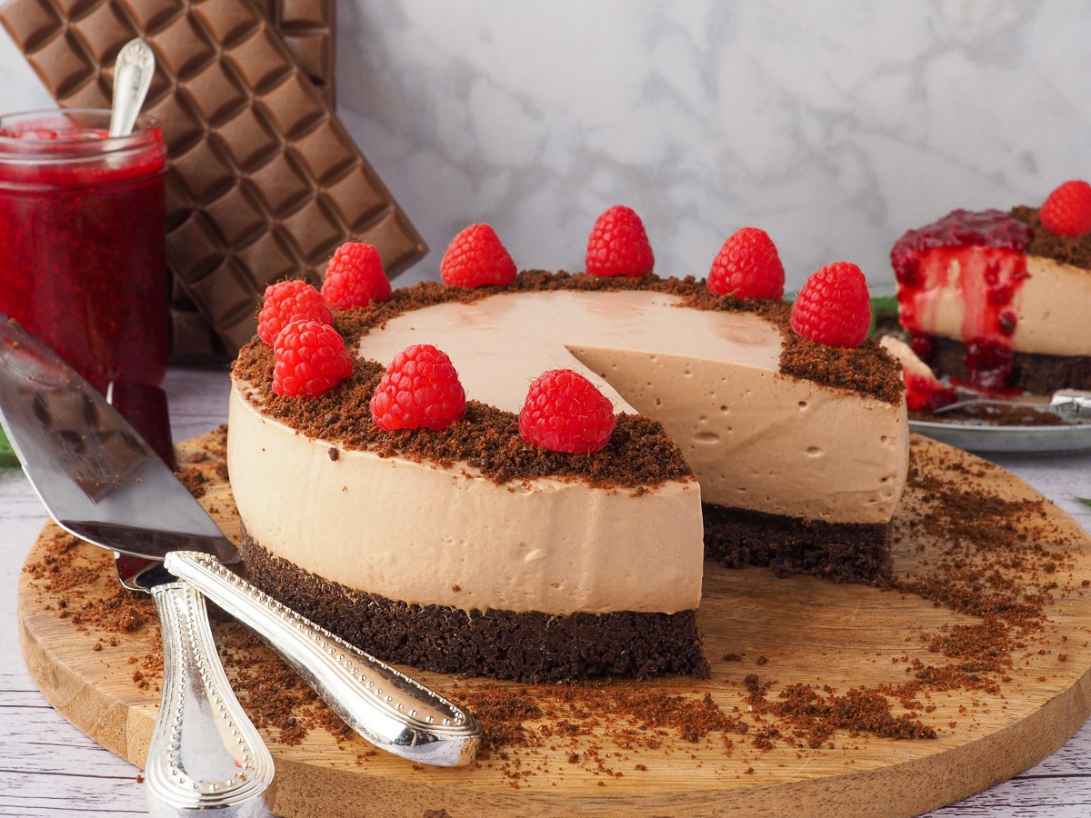 Cheesecake on a serving board, garnished with raspberries, with searving wear and slice of cheesecake, raspberry compote and blocks of chocolate in the background.