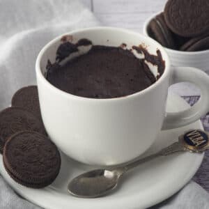 Close up mug cake on a saucer with a spoon and Oreos on the side and in the background.