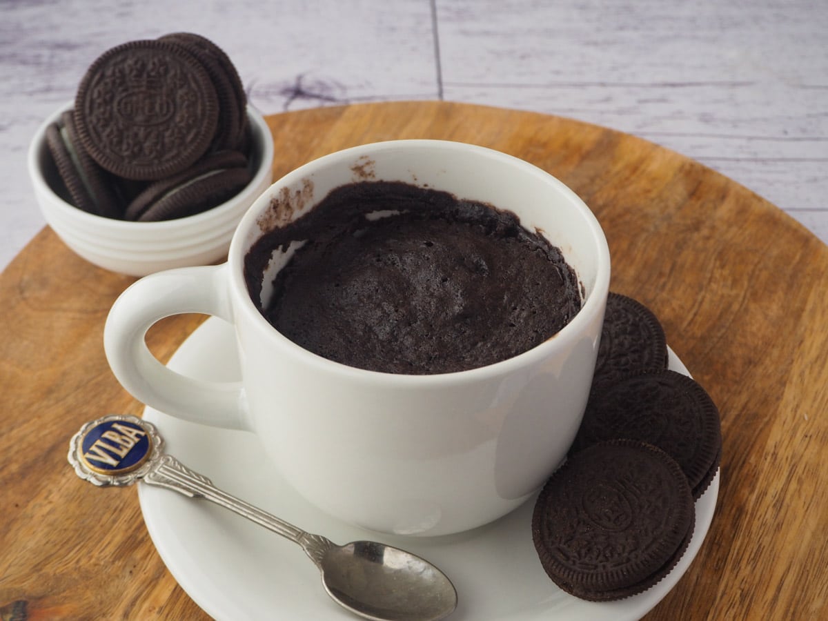 Mug cake on a saucer and board, with a spoon and Oreos on the side and in the background.