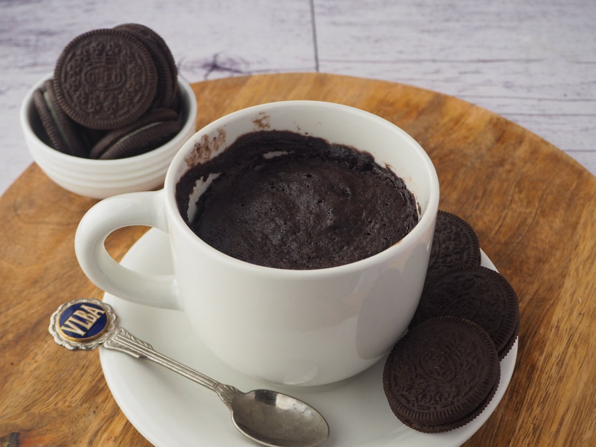 Oreo Mug Cake 15 Mug cake on a saucer and board, with a spoon and Oreos on the side and in the background.