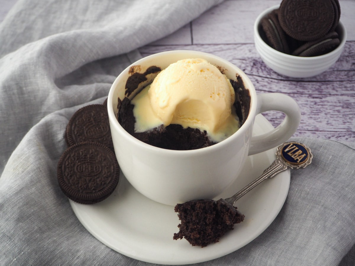Mug cake with a scoop of ice cream on a saucer with a spoon and Oreos on the side and in the background.