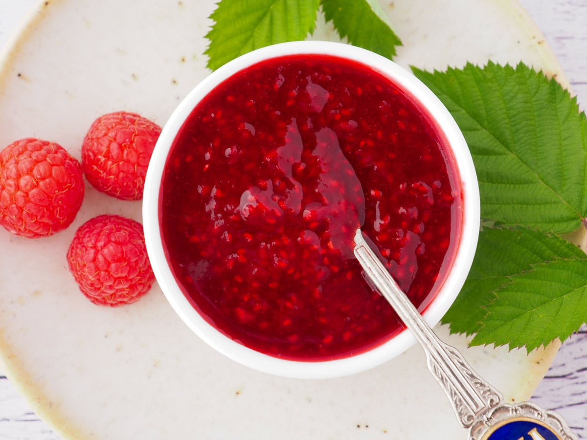 Top down view of compote in a serving bowl with a spoon, with fresh raspberries and leaves on the side.