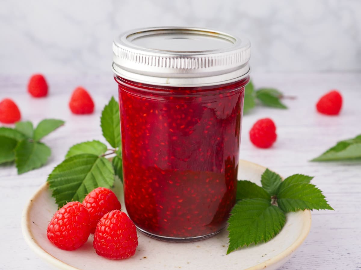 Raspberry Compote 2 Compote in a mason jar on a plate, with spoon and fresh raspberries and leaves.