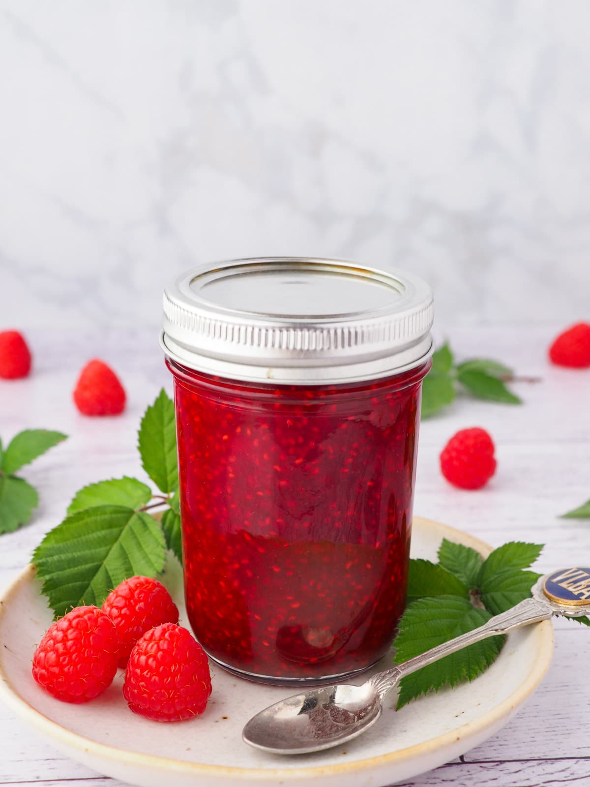Raspberry Compote 13 Compote in a mason jar on a plate, with spoon and fresh raspberries and leaves.