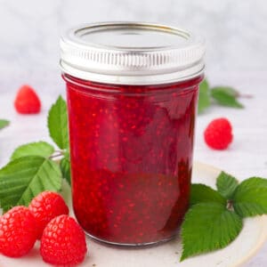 Raspberry Compote 11 Compote in a mason jar on a plate, with spoon and fresh raspberries and leaves.