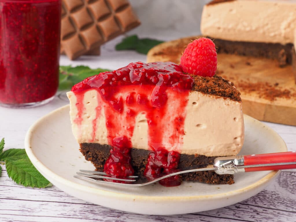 No bake chocolate cheesecake topped with raspberry compote on a plate with a fork, jar of raspberry compote and rest of cheesecake in the background.