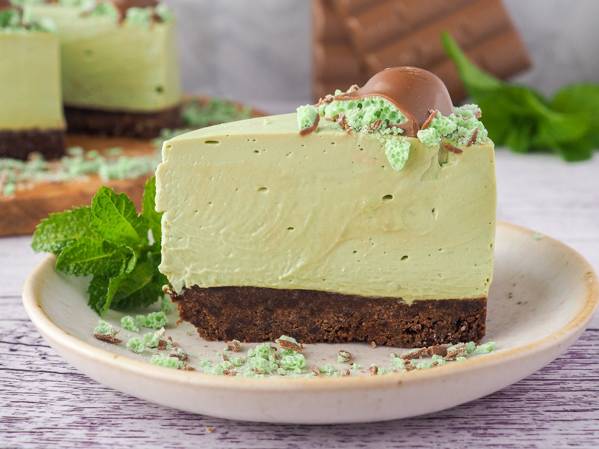 Slice of cheesecake on a plate, with cake and chocolate in the background.