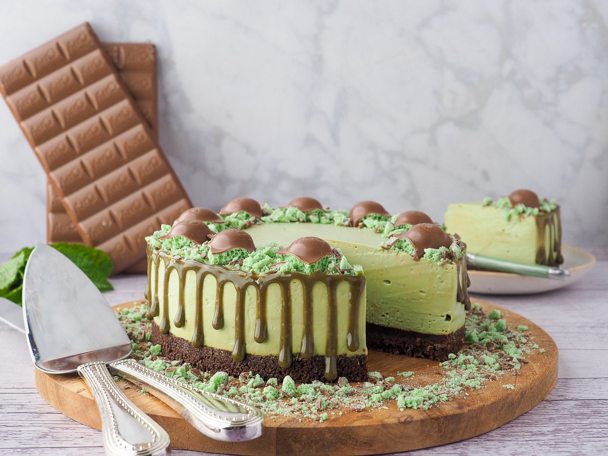 Cheesecake on a serving board, with serving wear, slice of cake and chocolate in the background.