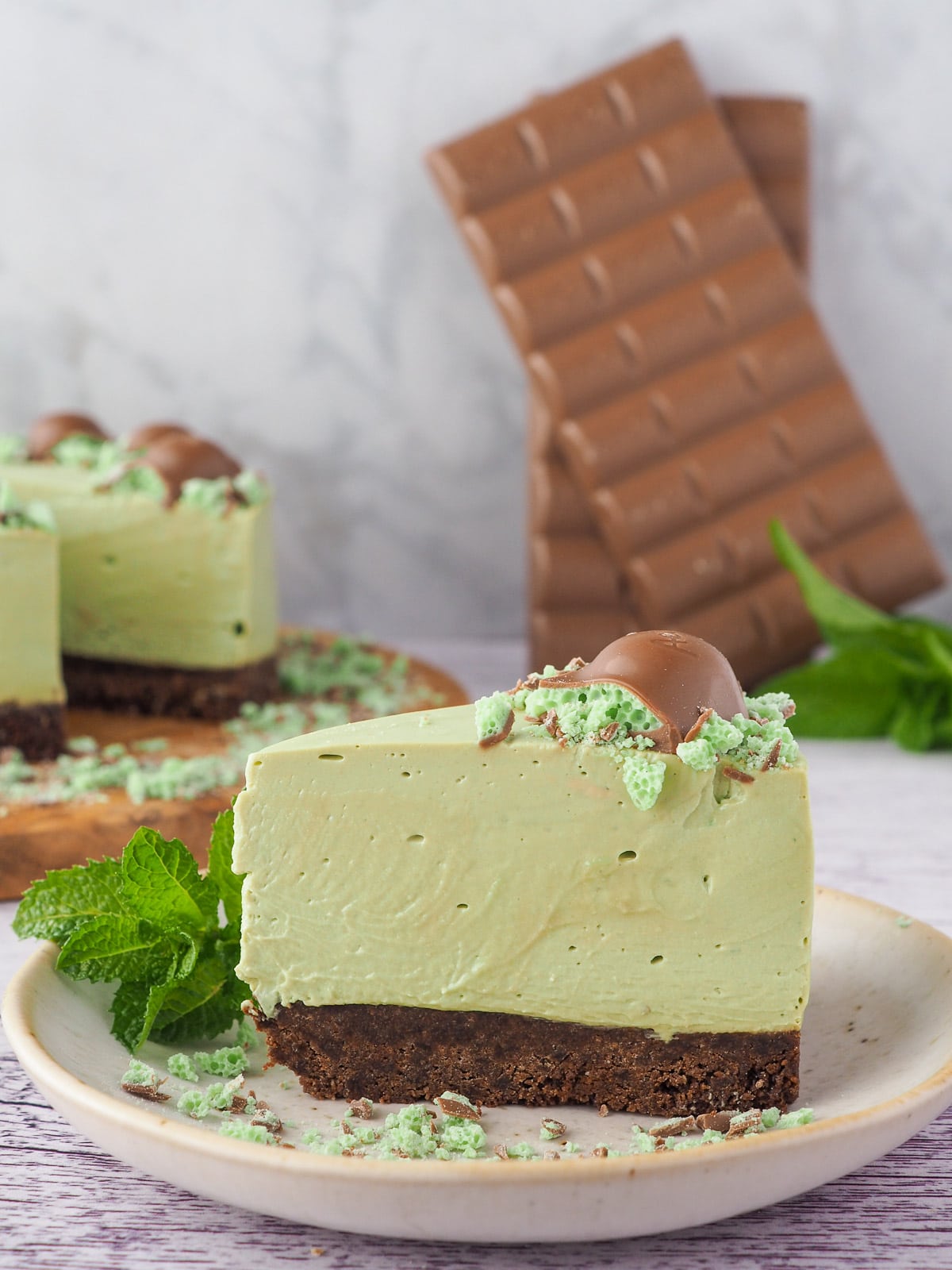 Aero Mint Cheesecake 12 Slice of cheesecake on a plate with cake and chocolate in the background.