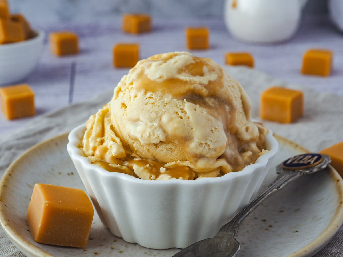 Ice cream with Bourbon butterscotch swirl, on a plate with a spoon, jug of butterscotch in the background and butterscotch candy scattered around it.