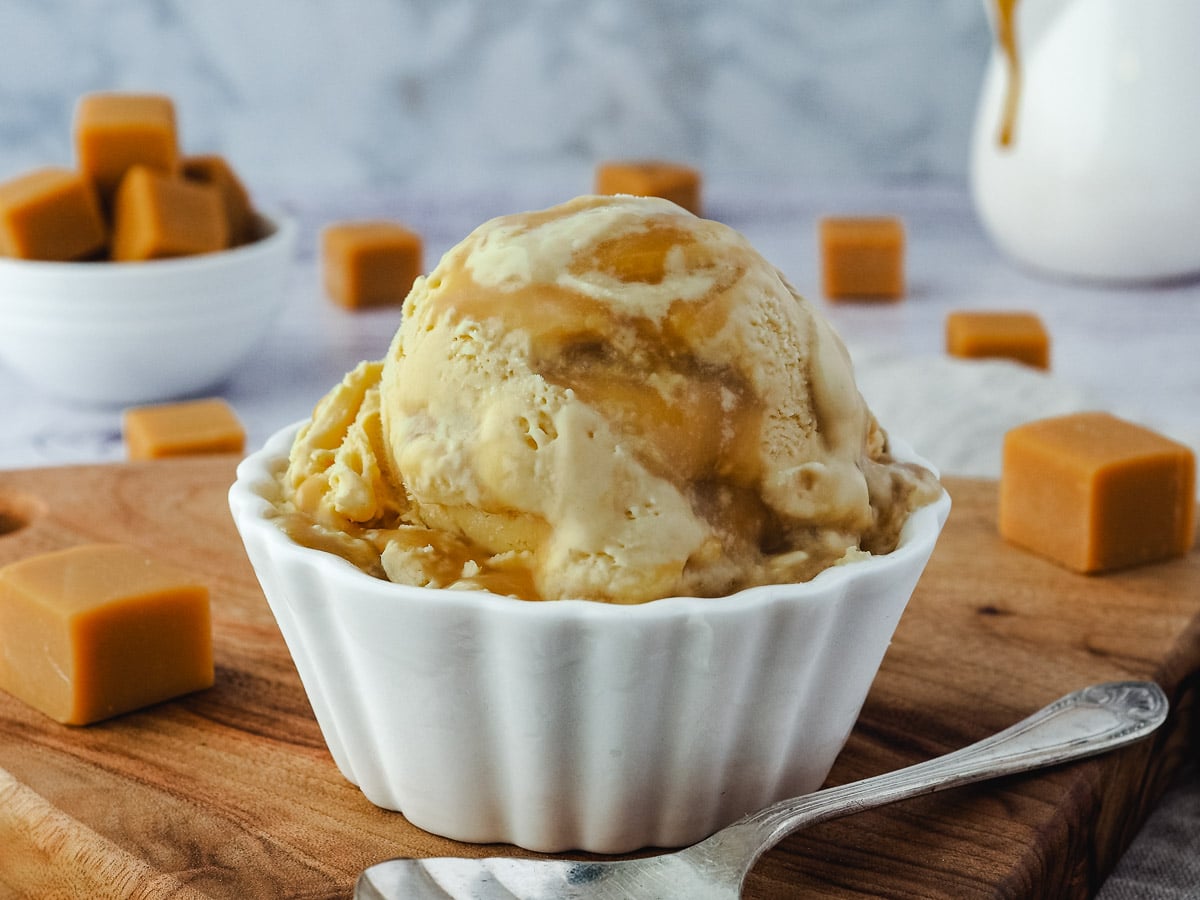 Ice cream with Bourbon butterscotch swirl, on a plate with a spoon, jug of butterscotch in the background and butterscotch candy scattered around it.