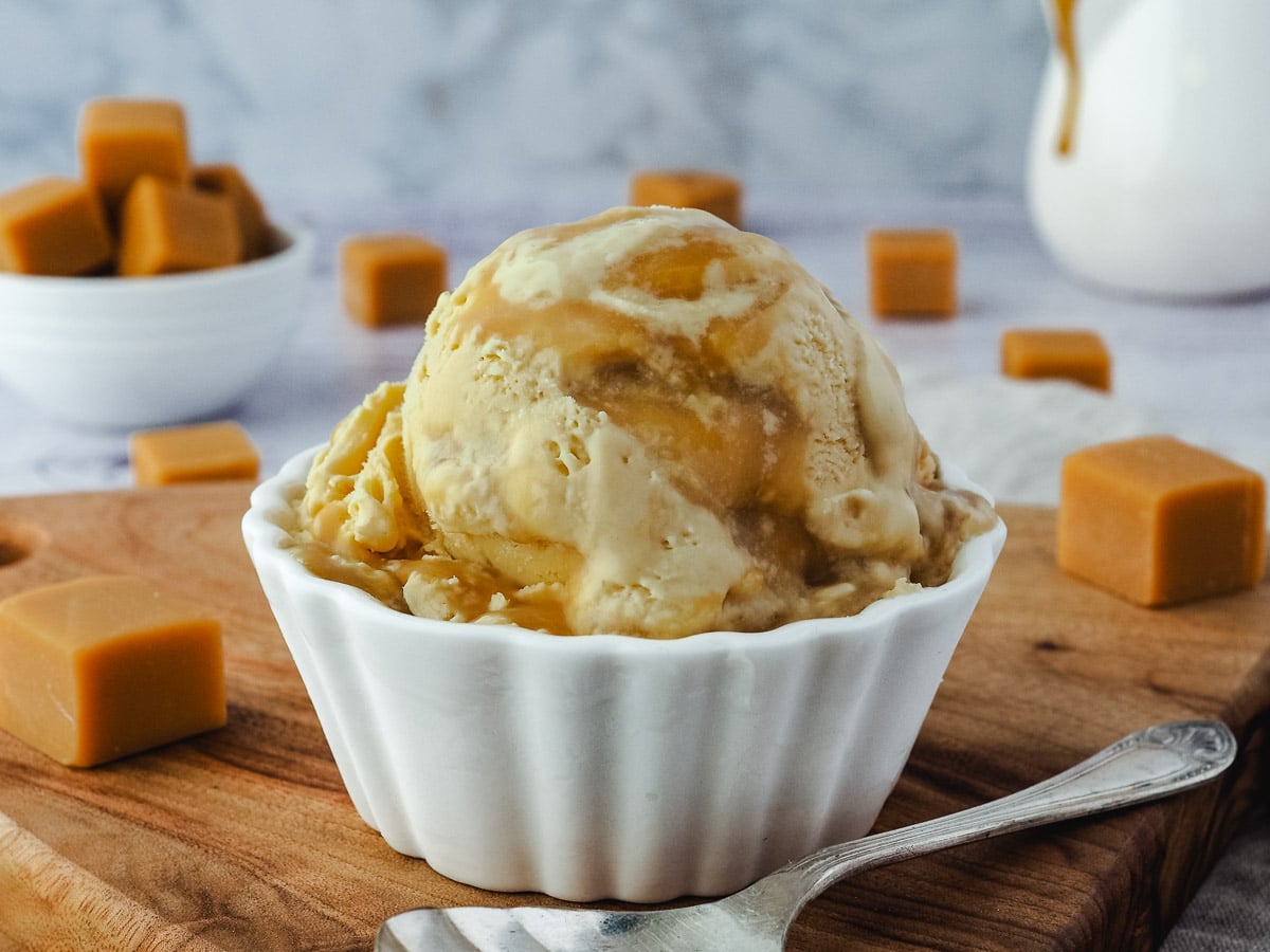 Butterscotch Ice Cream 9 Ice cream with Bourbon butterscotch swirl, on a plate with a spoon, jug of butterscotch in the background and butterscotch candy scattered around it.