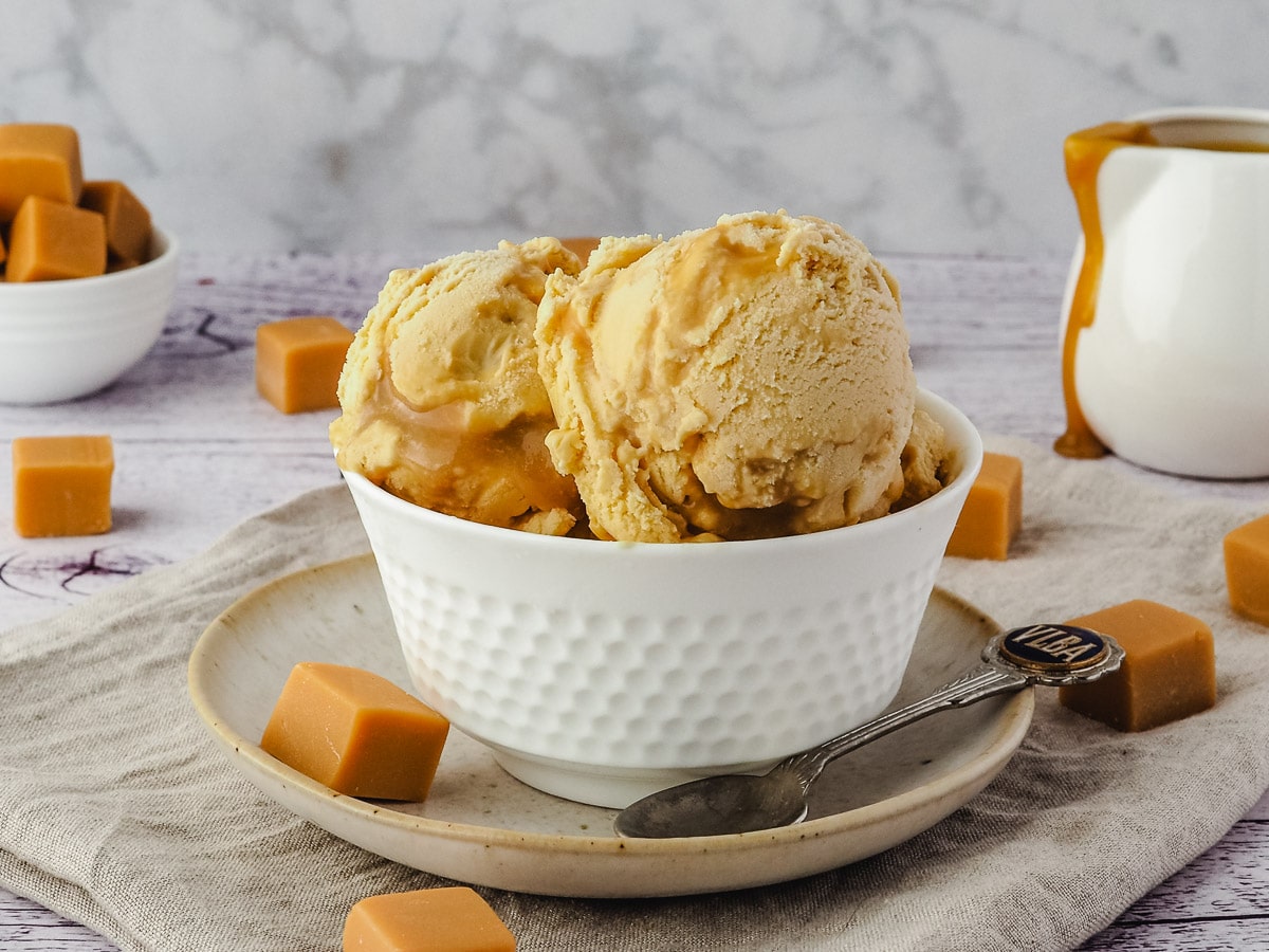 Ice cream with Bourbon butterscotch swirl, on a plate with a spoon, jug of butterscotch in the background and butterscotch candy scattered around it.