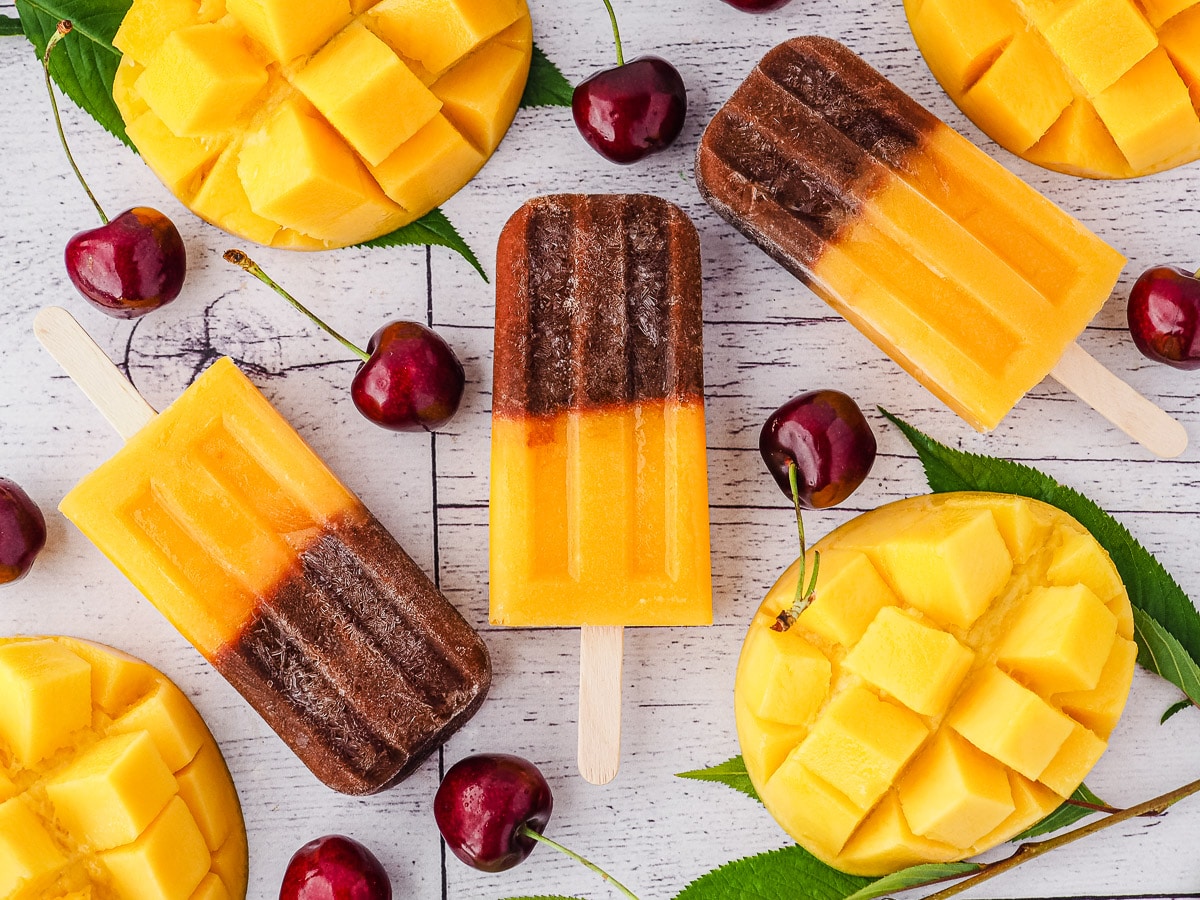 Cherry Mango Popsicles 2 Popsicles surrounded by fresh cherries and mango.