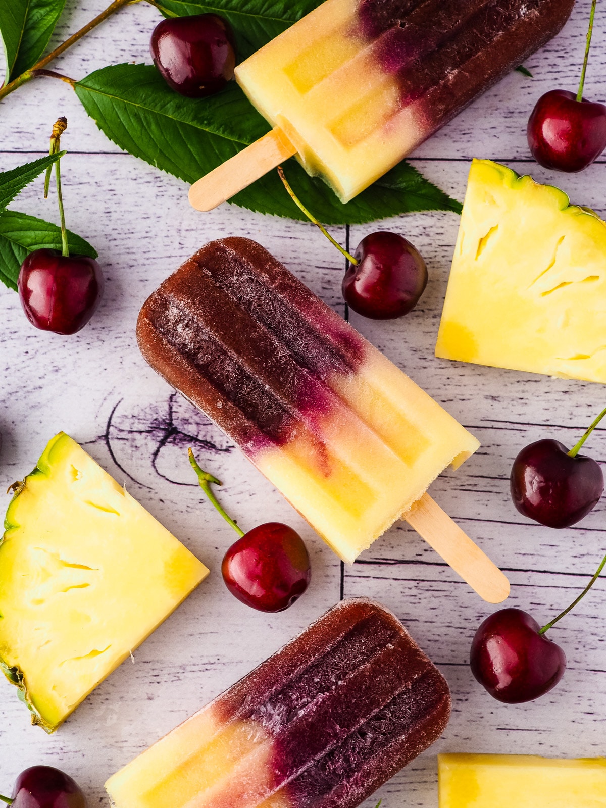 Popsicles surrounded by fresh pineapple, cherries and cherry leaves.