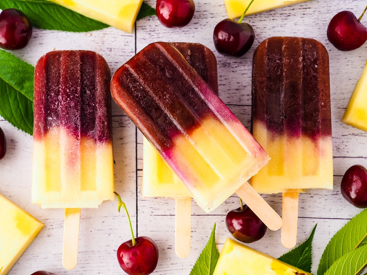 Stack of popsicles surrounded by fresh pineapple, cherries and cherry leaves.