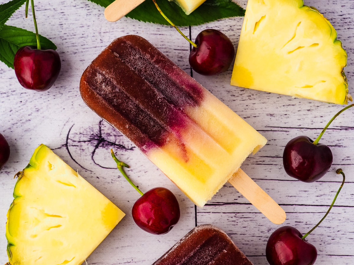 Close up popsicle surrounded by fresh pineapple, cherries and cherry leaves.