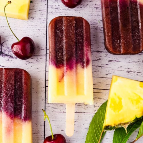 Close up popsicles surrounded by fresh pineapple, cherries and cherry leaves.