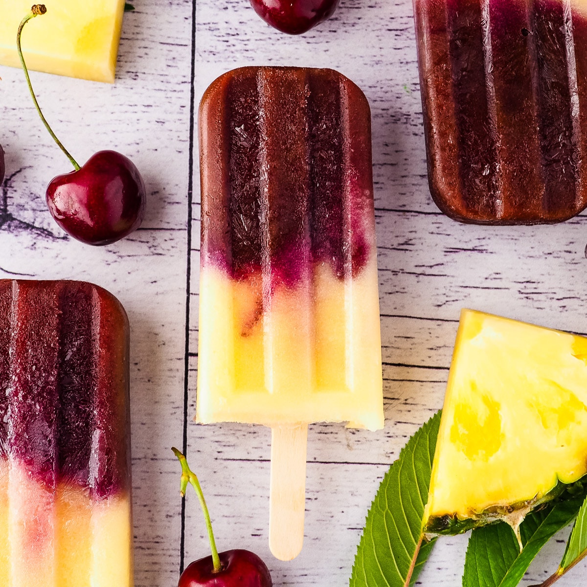 Close up popsicles surrounded by fresh pineapple, cherries and cherry leaves.