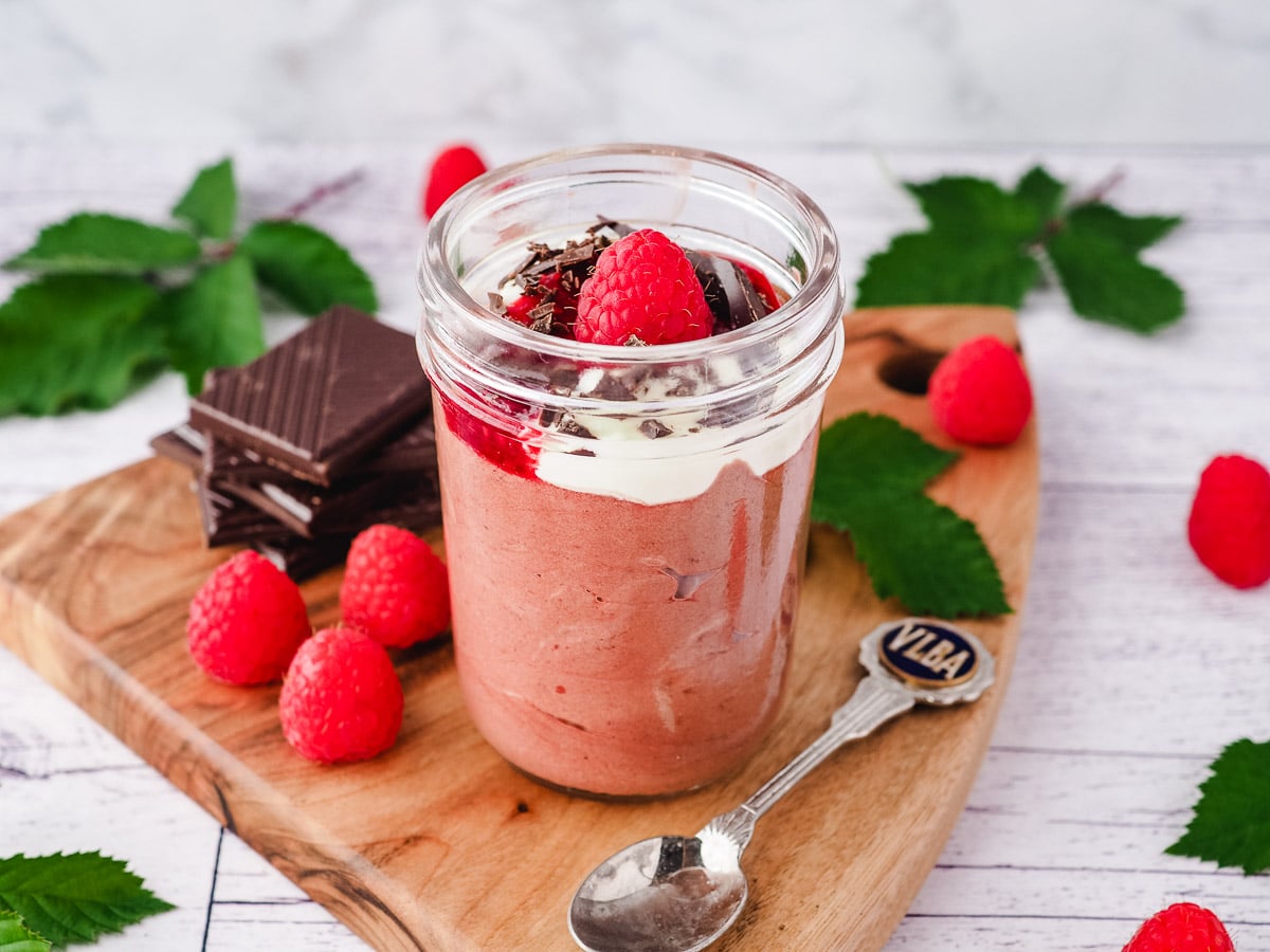 Mousse in mason jar, garnished with cream, raspberry compote, chocolate and fresh raspberries, with a spoon on the side, surrounded by fresh raspberries and leaves and a stack of chocolate.