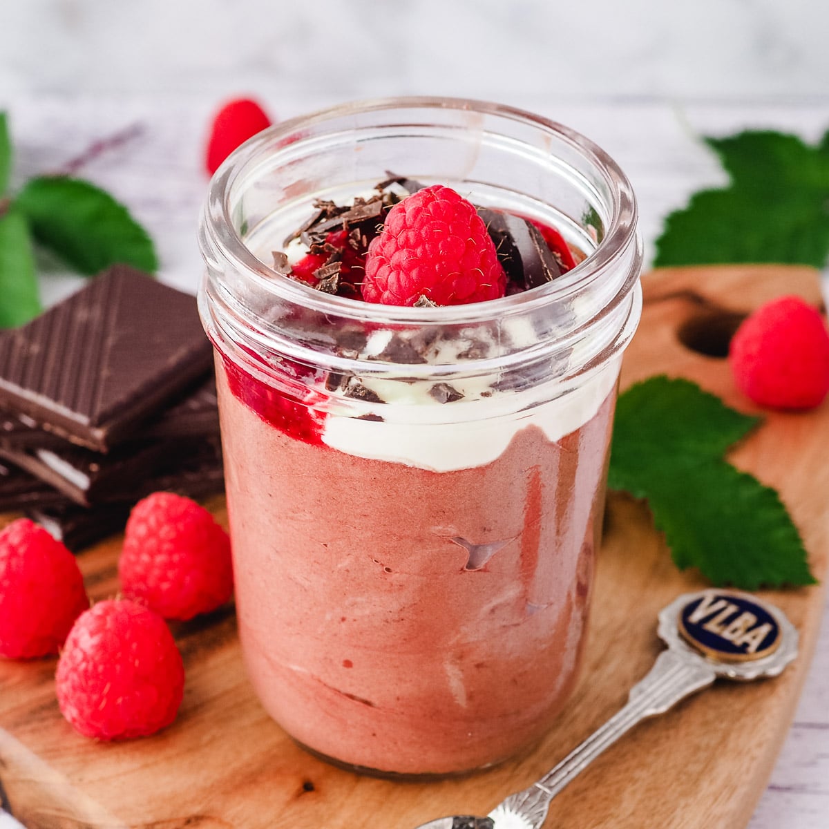 Close up mousse in mason jar, garnished with cream, raspberry compote, chocolate and fresh raspberries, with a spoon on the side, surrounded by fresh raspberries and leaves and a stack of chocolate.