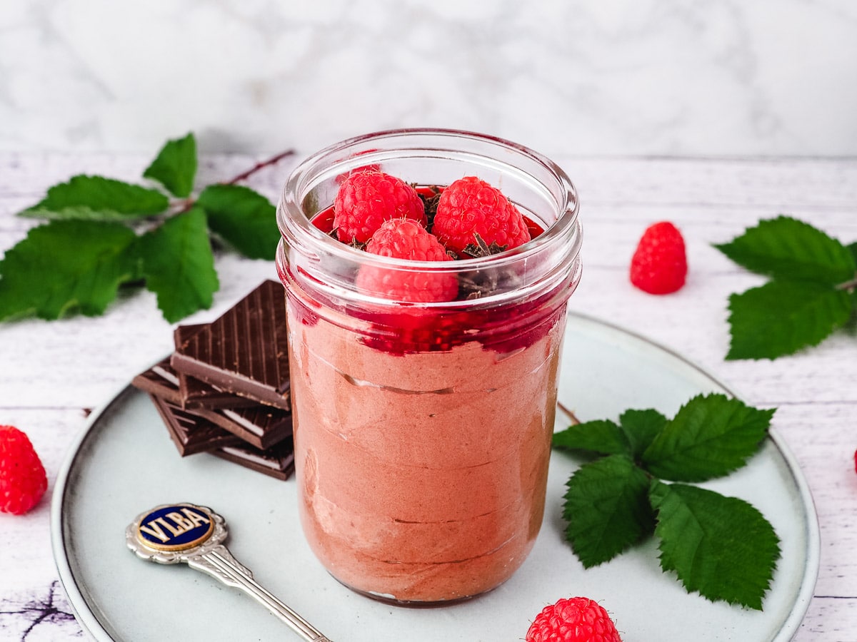 Mousse in mason jar, garnished with raspberry compote, chocolate and fresh raspberries, with a spoon on the side, surrounded by fresh raspberries and leaves and a stack of chocolate.