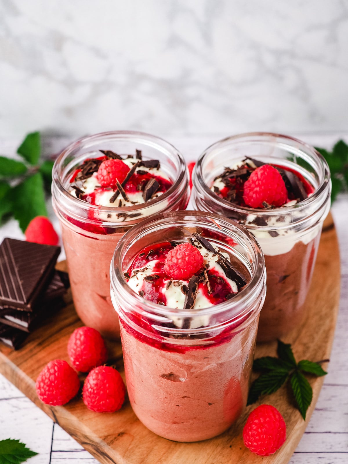 Group of three mousse in mason jars, garnished with cream, raspberry compote, chocolate and fresh raspberries, with a spoon on the side, surrounded by fresh raspberries and leaves and a stack of chocolate.