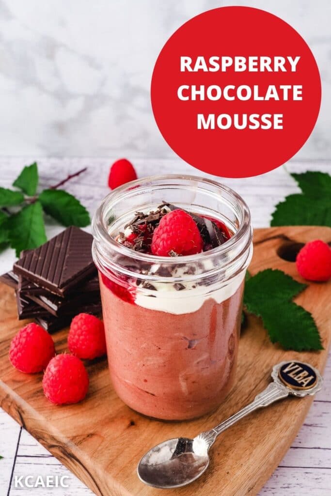 Mousse in mason jar, garnished with cream, raspberry compote, chocolate and fresh raspberries, with a spoon on the side, surrounded by fresh raspberries and leaves and a stack of chocolate, with text overlay, raspberry chocolate mousse and KCAEIC.