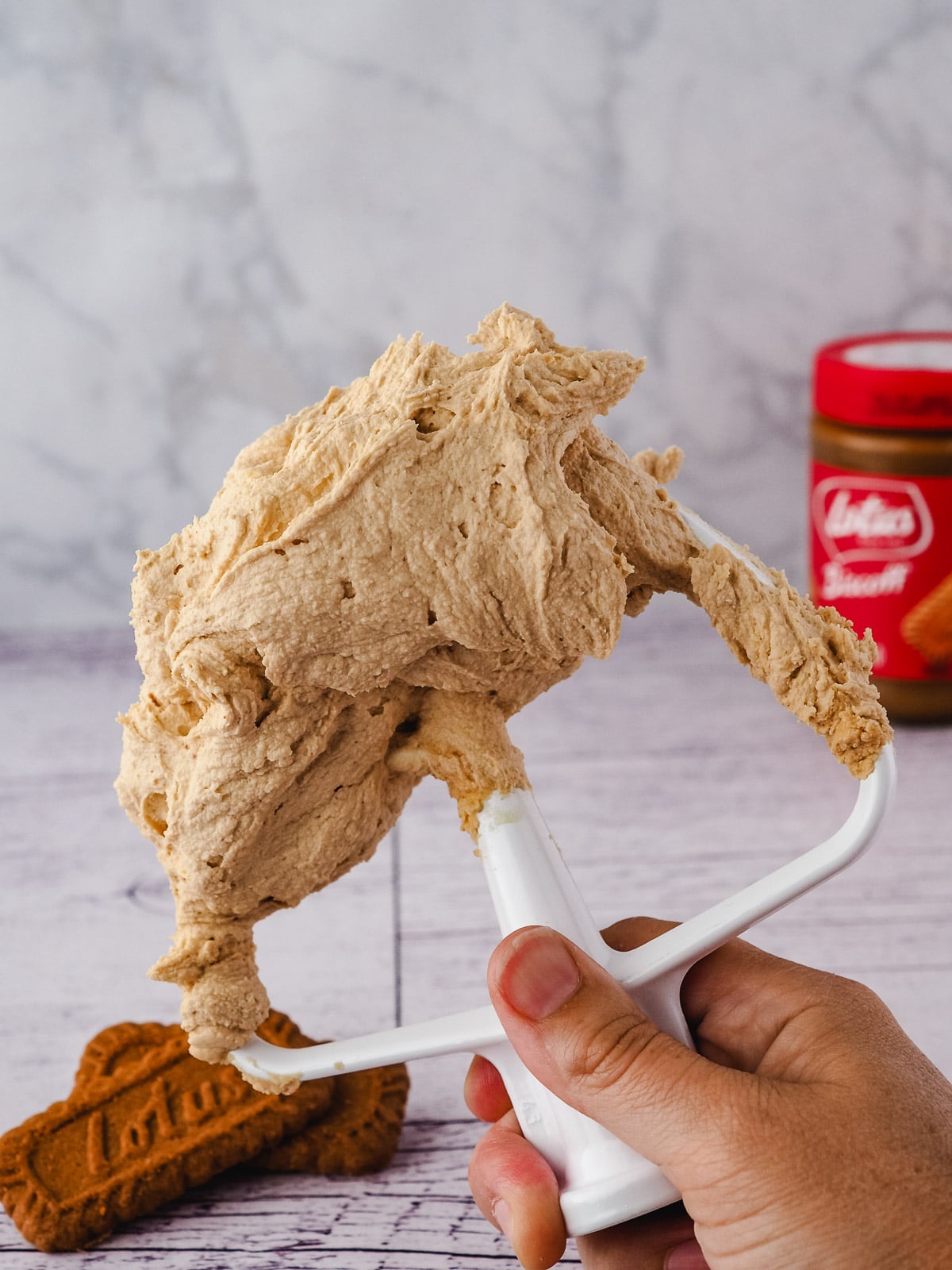 Hand holding a beater with Biscoff buttercream frosting, with biscuits and jar of spread in the background.