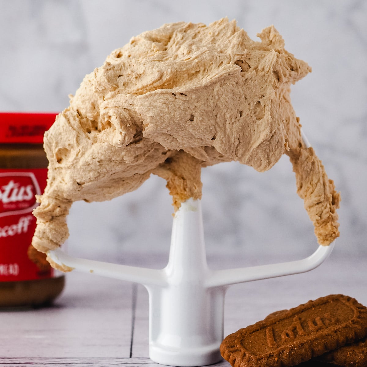 Close up buttercream on beater with biscuits to the side and jar of spread in background.