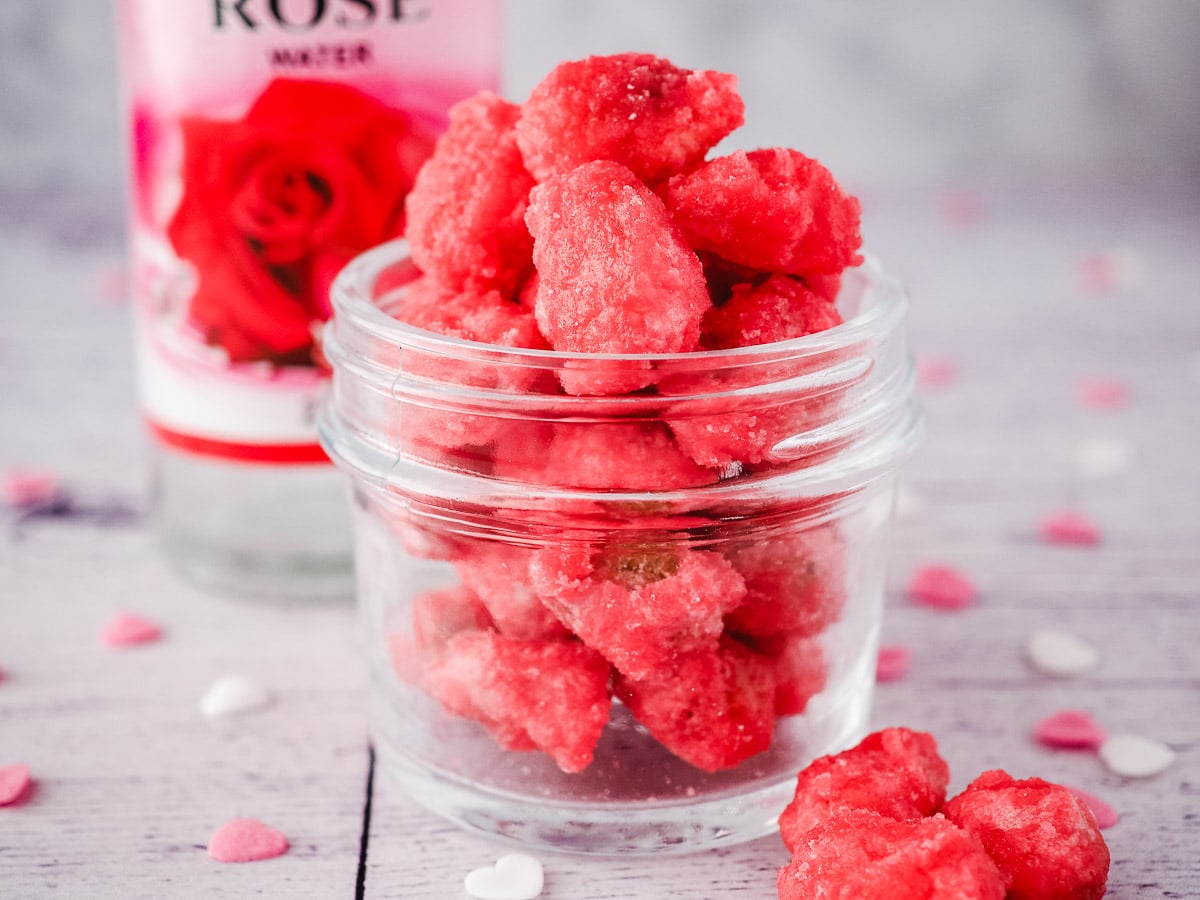 Small mason jar filled with pralines, surrounded by hear sprinkles, with rose water in the background.