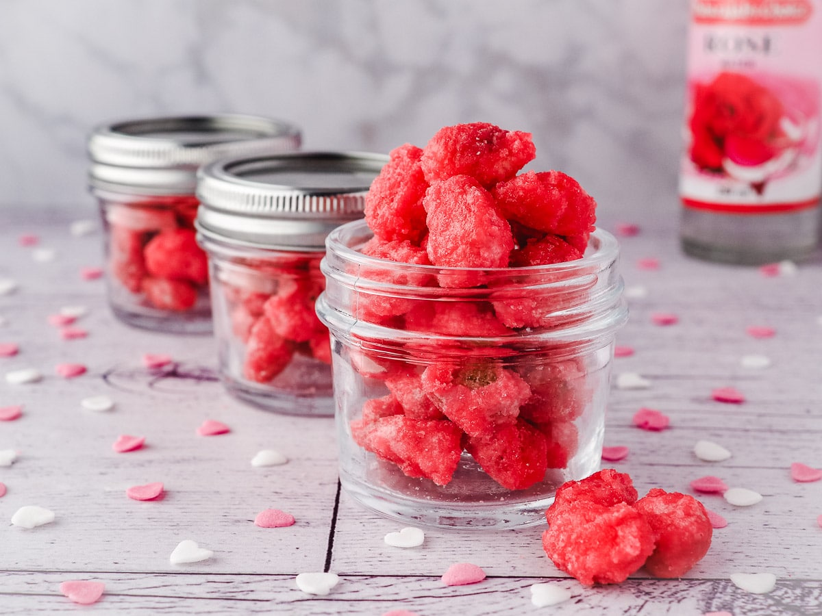 Row of small mason jars filled with pralines, surrounded by hear sprinkles, with rose water in the background.