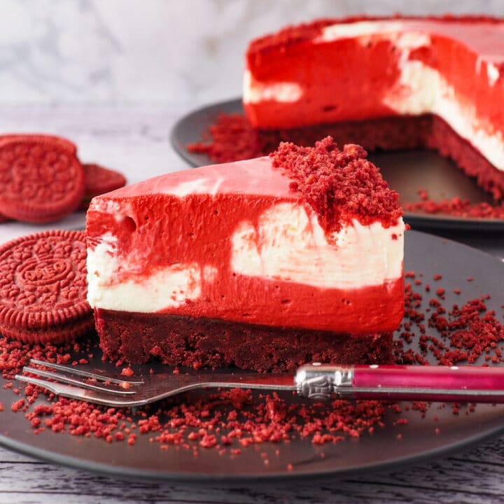 Cheesecake on a plate with a fork and crumbs, with red velvet Oreos and rest of cake in the background.