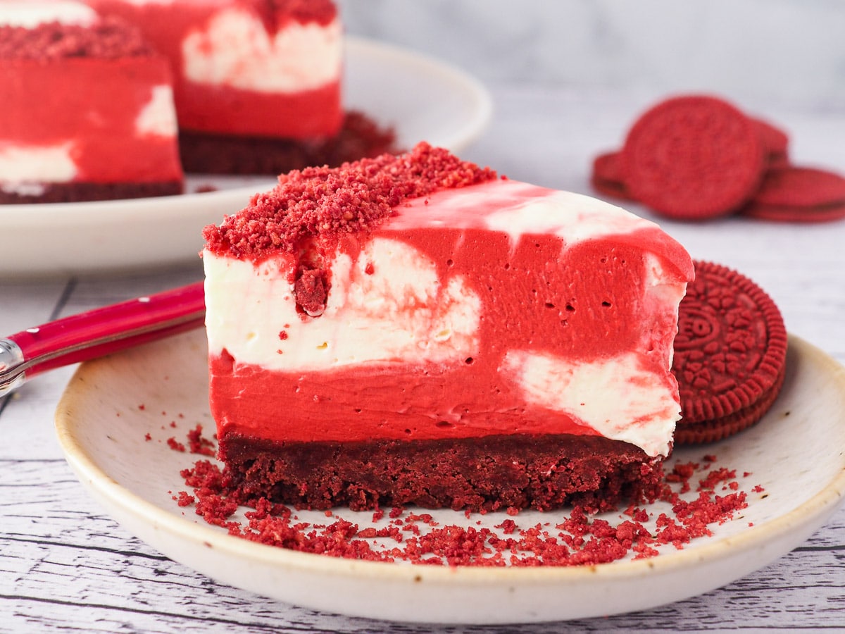 Slice of cheesecake on a plate with a fork and crumbs, with red velvet Oreos and rest of cake in the background.