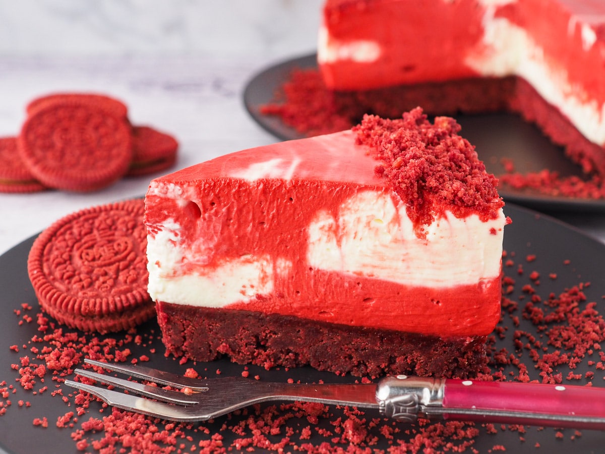 Red Velvet Oreo Cheesecake 2 Slice of cheesecake on a plate with a fork and crumbs, with red velvet Oreos and rest of cake in the background.