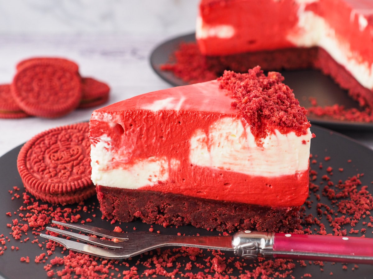 Slice of cheesecake on a plate with a fork and crumbs, with red velvet Oreos and rest of cake in the background.