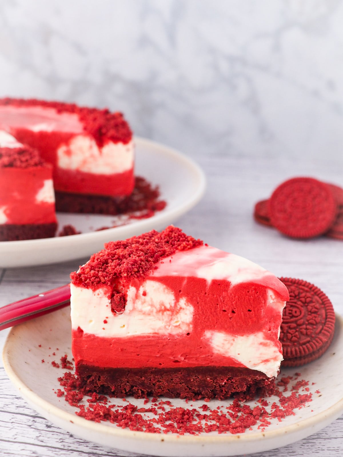 Cheesecake on a plate with a fork and crumbs, with red velvet Oreos and rest of cake in the background.