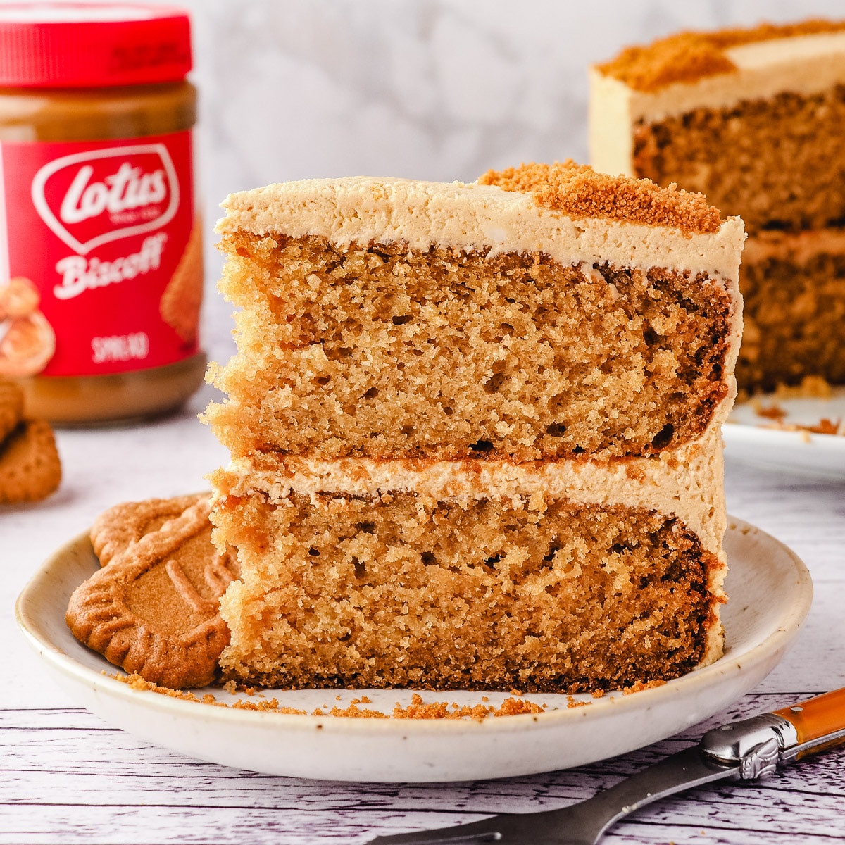 Close up cake on a plate with fork and rest of cake, jar of Biscoff spread and Biscoff biscuits in background.